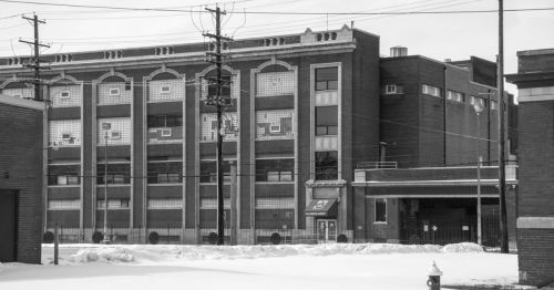 A large, historic brick building with multiple windows, surrounded by snow and utility poles, in black and white.
