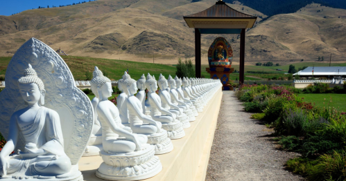 A row of white Buddha statues lined along a path, with mountains and a colorful shrine in the background.