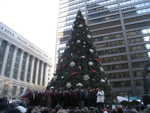 A large decorated Christmas tree stands in a city square, with a choir performing in front of it. Tall buildings surround the scene.
