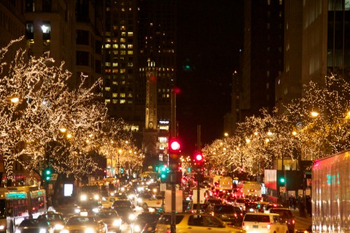 A bustling city street at night, adorned with twinkling lights on trees, with traffic and illuminated buildings in the background.