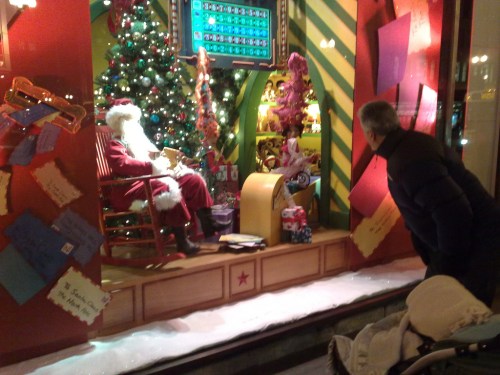 A festive window display featuring Santa Claus, a Christmas tree, and a person admiring the scene.