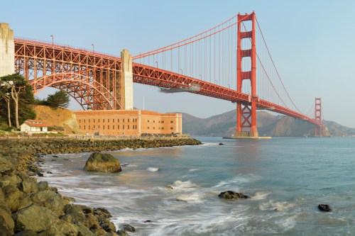 Golden Gate Bridge spans over water, with rocky shore and historic buildings in the foreground, under a clear sky.