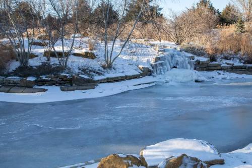 A frozen pond with a small waterfall, surrounded by snow-covered trees and rocks.