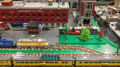 A detailed model train setup featuring buildings, tracks, trees, and vehicles in a vibrant, colorful scene.