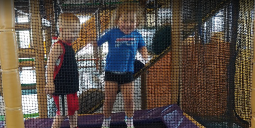 Two young children play on a trampoline inside a play structure, surrounded by safety netting.