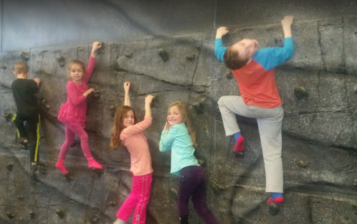 Five children climbing a rock wall indoors, smiling and enjoying their activity.