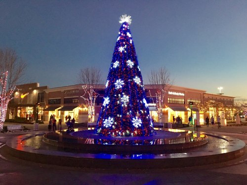 A brightly lit Christmas tree adorned with blue and white decorations stands in a plaza at dusk, surrounded by shops.