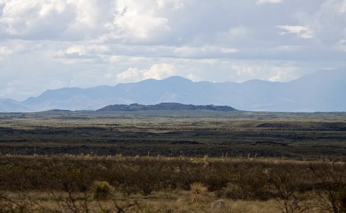 A vast landscape with rolling hills and distant mountains under a cloudy sky. Sparse vegetation in the foreground.