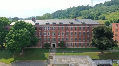 A large, red-brick building with multiple windows, surrounded by trees and greenery, set against a hillside.