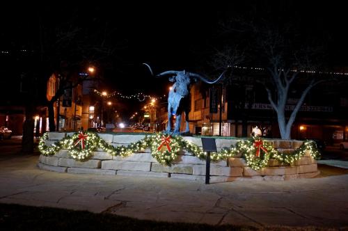 A longhorn statue adorned with festive lights and greenery, set in a town square at night.