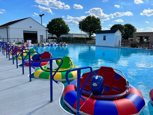 Colorful bumper boats lined up at the edge of a clear blue pool under a sunny sky with fluffy clouds.