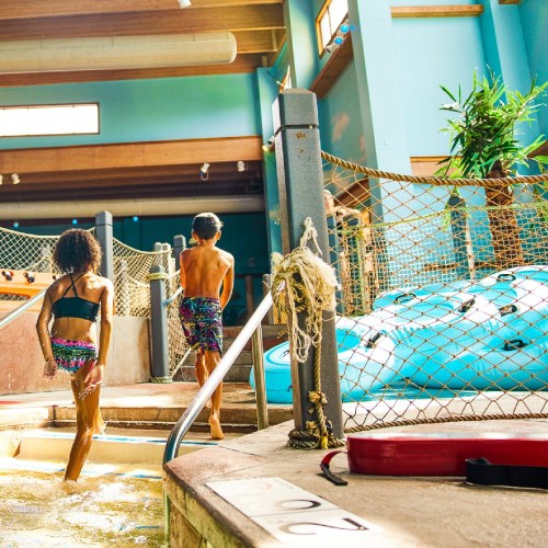 Two children in swimwear walk towards a water slide in a brightly lit indoor water park.
