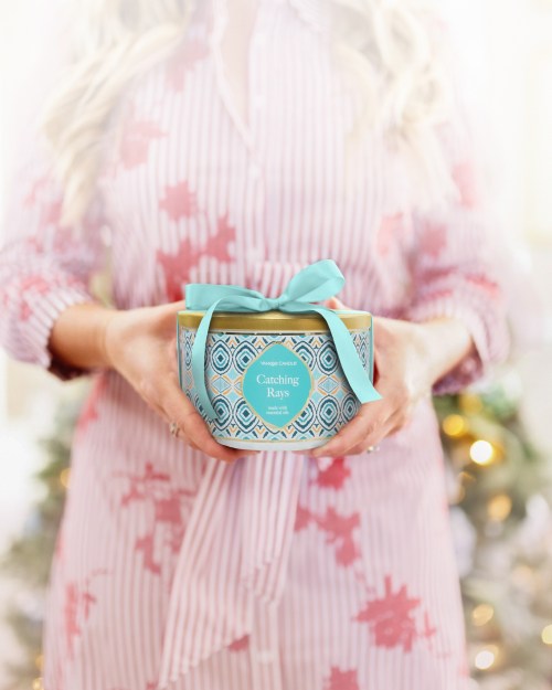A person holds a decorative candle labeled "Catching Rays," wrapped with a light blue ribbon, in a festive setting.