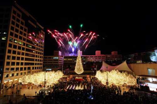 A festive scene with a large Christmas tree, twinkling lights, and fireworks illuminating the night sky above a crowd.