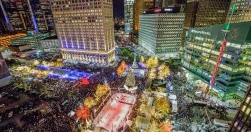 Aerial view of a bustling city square at night, filled with people, festive lights, and an ice skating rink.
