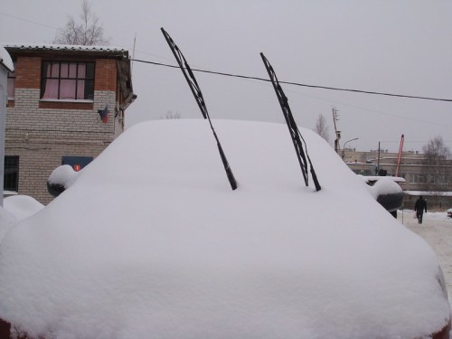 A car covered in snow with its windshield wipers sticking up, set against a snowy urban backdrop.