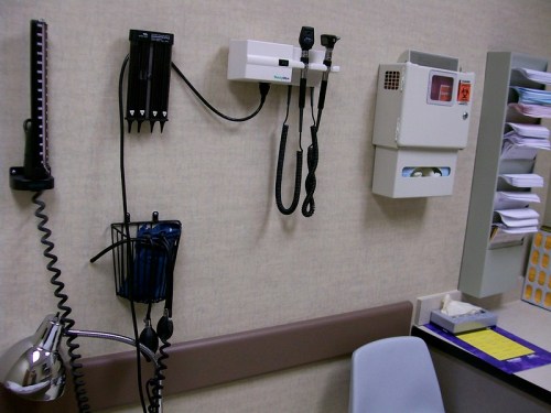 Medical examination room with diagnostic tools on the wall, including an otoscope and sphygmomanometer.
