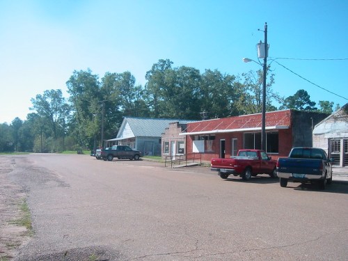 A quiet rural street with old buildings and parked cars under a clear blue sky. Trees line the background.