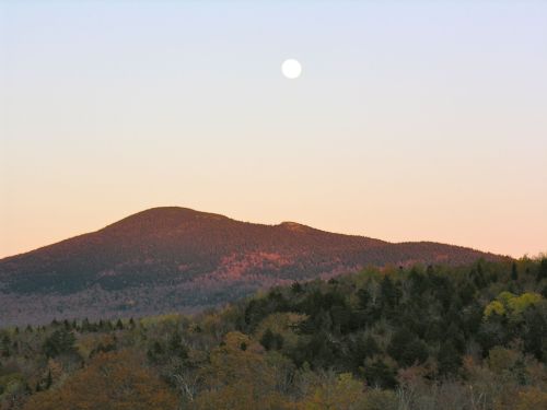 A serene mountain landscape at dusk, with a full moon rising above the peaks and colorful autumn foliage below.