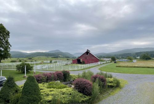 A red barn surrounded by green fields and mountains under a cloudy sky, with a white fence and landscaped garden in the foreground.