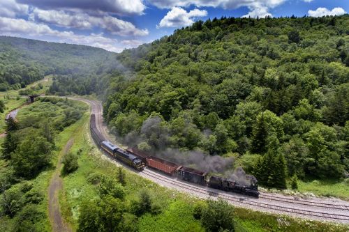 A steam train travels along a winding track through lush green hills under a partly cloudy sky.