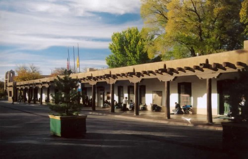 A sunlit plaza with adobe-style architecture, featuring trees and people sitting under a covered walkway.