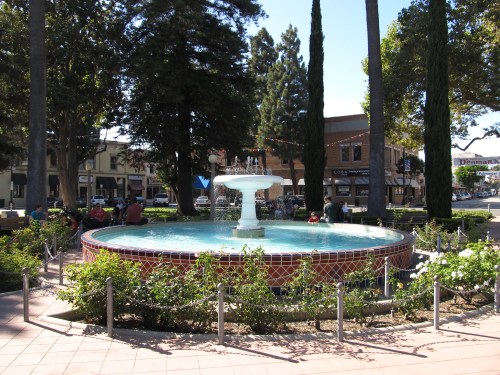A circular fountain surrounded by trees and shops, with people sitting nearby in a sunny park setting.