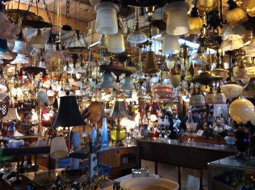 A cluttered antique shop filled with various hanging lamps and light fixtures, creating a warm, inviting atmosphere.