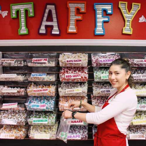 A smiling woman in an apron fills a bag with colorful taffy from labeled bins in a candy shop.