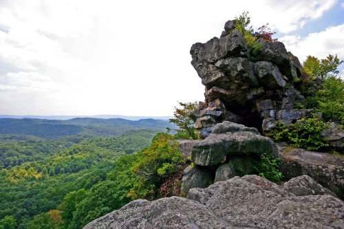 A rocky outcrop overlooks a lush green valley and distant mountains under a partly cloudy sky.