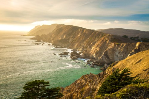 A scenic coastal view with rugged cliffs, gentle waves, and a golden sunset illuminating the landscape.