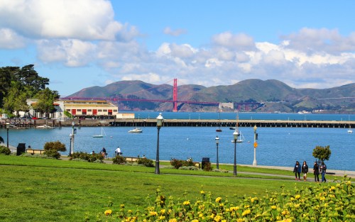 A scenic view of the Golden Gate Bridge from a park, with water, boats, and people walking along the shore.