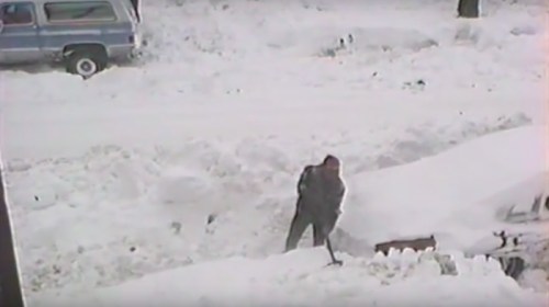 A person shoveling snow from a driveway, surrounded by deep snow and parked vehicles in a winter scene.
