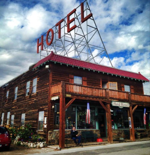 A rustic wooden hotel with a large "HOTEL" sign, featuring a porch and flowers, under a cloudy sky.