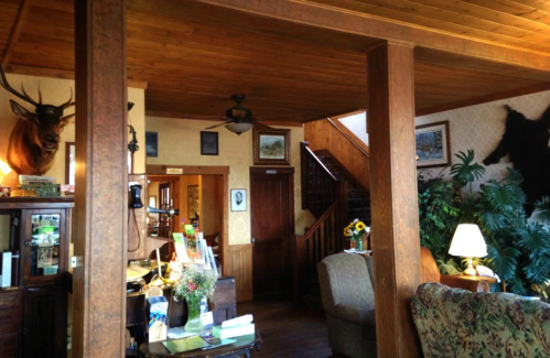 Cozy interior of a rustic lodge with wooden beams, mounted animal heads, and a staircase in the background.