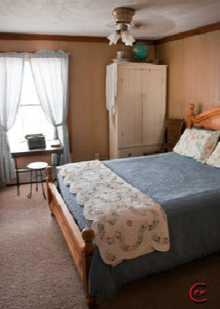 Cozy bedroom with a wooden bed, blue bedding, vintage curtains, and a small table by the window.