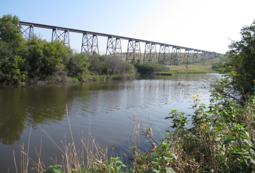 A tall railway bridge spans over a calm river, surrounded by lush greenery and open fields under a clear blue sky.