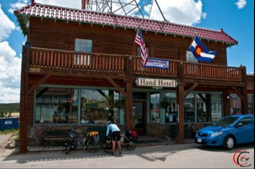 A wooden hotel with a red roof, flags, and a cyclist near parked bikes in a small town setting.