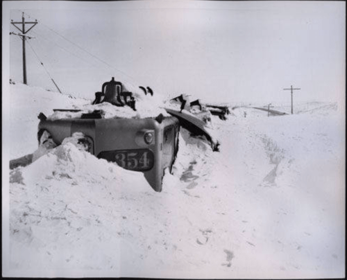 A snow-covered train partially buried in a blizzard, with power lines visible in the background.