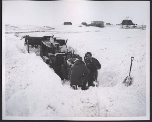 A group of people working to clear snow around a vehicle in a snowy landscape with distant buildings.