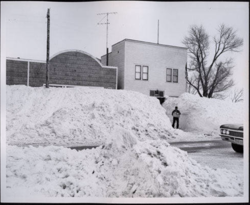 A person stands beside a large snowbank near a building, with more snow piled up in the foreground and background.