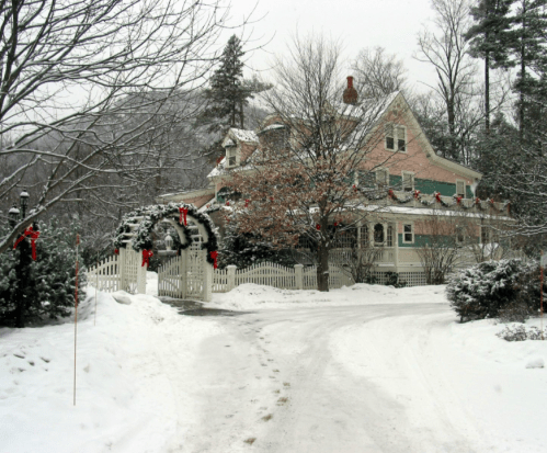 A snowy scene of a charming house decorated for the holidays, with wreaths and red bows on the porch and trees.