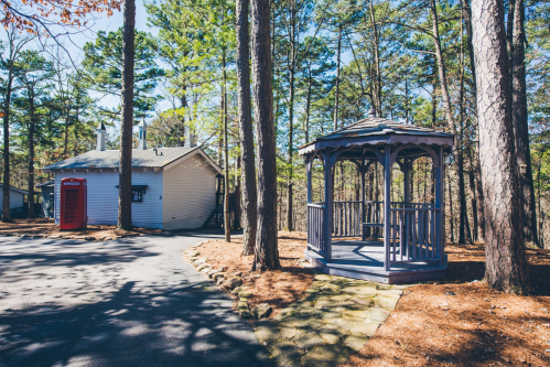 A wooden gazebo beside a white house, surrounded by tall trees and a paved path in a sunny forest setting.