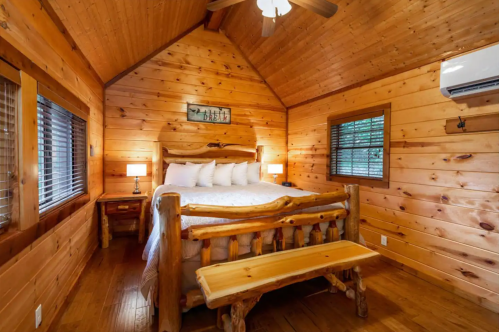 Cozy wooden cabin interior featuring a bed, nightstands, and a bench under a ceiling fan. Warm, rustic decor.