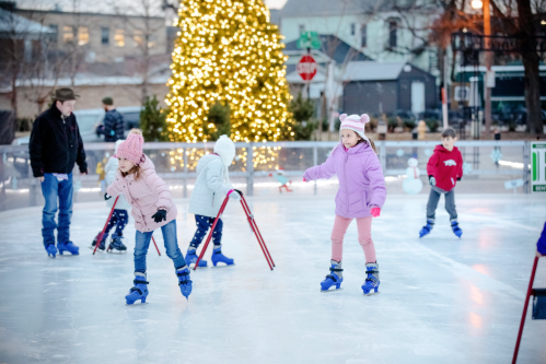 Children ice skating on a rink decorated with a Christmas tree, with adults supervising in a festive outdoor setting.