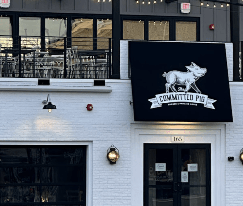 Sign for "The Committed Pig" restaurant featuring a pig graphic, with a modern building and outdoor seating area.
