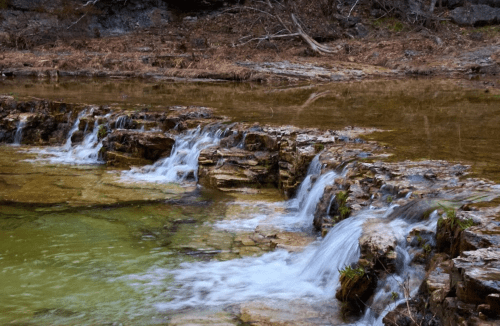 A serene stream flows over rocky formations, surrounded by earthy tones and calm water.