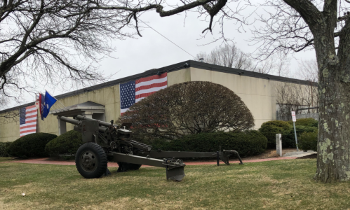 A military cannon displayed on grass in front of a building with American and state flags. Trees are in the foreground.