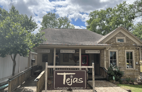 A rustic building with a sign reading "Tejas Chocolate + Barbecue" surrounded by trees and a blue sky.
