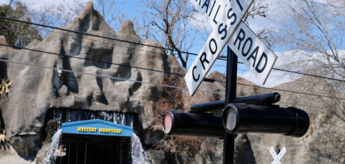 A railroad crossing sign in front of a mountain-themed amusement attraction with a waterfall.